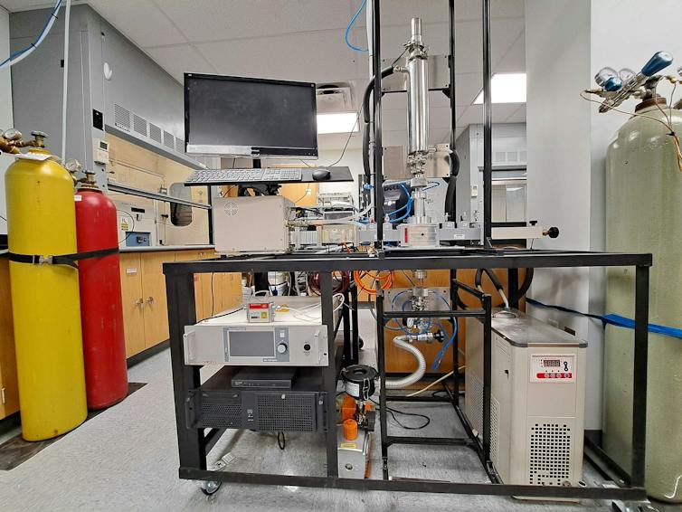Photo of a chemistry lab space with colorful gas bottles. At the center of the image is a microwave reactor connected by a waveguide to a microwave source.